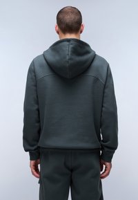Mørkegrøn hættebluse med struktureret overflade og hætte, der har et enkelt design og tætsiddende manchetter, parret med matchende mørke svedbukser.