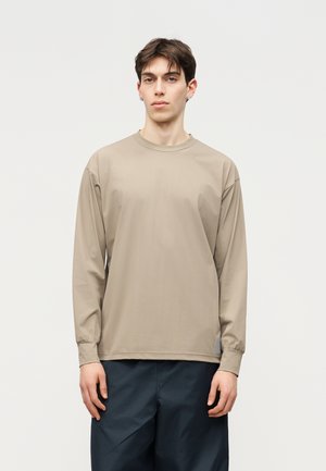 Jonge man met kort bruin haar, gekleed in een beige shirt met lange mouwen en donkere broek, staand tegen een effen lichte achtergrond.