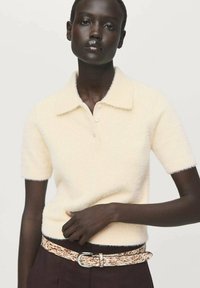 Modello che indossa una polo a maniche corte di colore crema in maglia fluffy con bottoni, abbinata a pantaloni scuri e una cintura bianca e marrone con texture.