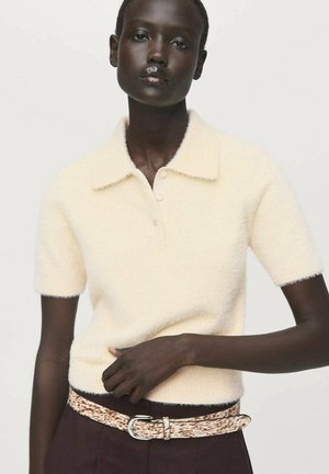 Modelo vistiendo una camiseta de polo de punto difuso en crema con mangas cortas y botones, combinada con pantalones oscuros y un cinturón texturizado en blanco y marrón.