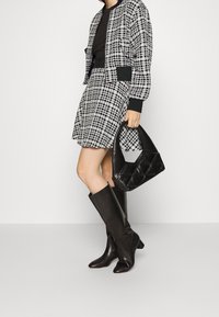 Veste bomber à carreaux et jupe assortie en noir et blanc, associées à des bottes en cuir noir montant jusqu'aux genoux et à un sac noir matelassé.
