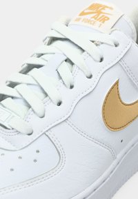 Bijele kožne tennisice Nike Air Force 1 sa zlatnim Swoosh-om, ravnim vezicama i teksturiranom površinom. Imaju pjenasti međusol i oznaku na jeziku.