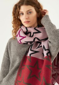 Écharpe tricotée en rose clair avec des étoiles noires, présentant une partie inférieure rouge et des bords frangés. Portée par-dessus un pull gris.