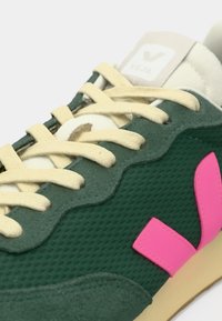 Πράσινο αθλητικό παπούτσι από δίχτυ με κυματιστές επενδύσεις από σουέτ, εκρού κορδόνια, ροζ λογότυπο στη πλευρά και μπεζ ελαστική σόλα.