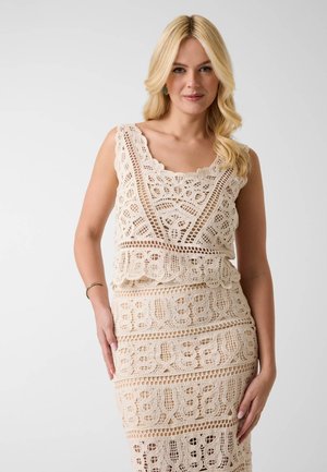 Mujer rubia con un vestido de ganchillo sin mangas de color crema con patrones florales y geométricos, posando sobre un fondo liso.