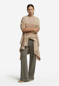 Beige stickad cardigan över en beige polotröja, i kombination med strukturerade grå vida byxor. Sneaker kompletterar outfiten. Tydlig bakgrund.