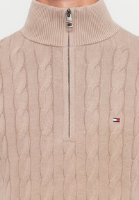Beige kabelgebreide trui met hoge geribbelde kraag, halve rits aan de voorkant en klein rood, wit en marineblauw logo op de borst.