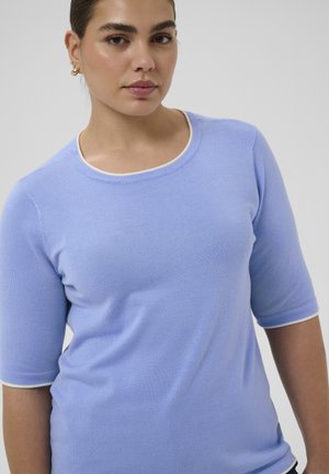 Kvinde med mørkt hår bundet tilbage iført en lyseblå kortærmet sweater med hvide kanter, set forfra mod en ensfarvet baggrund.