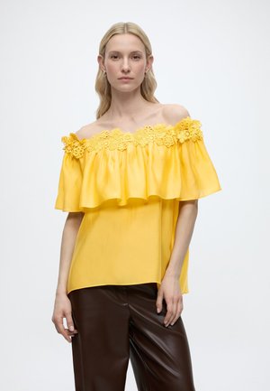 Kvinde iført gul off-shoulder top med blomstret blonderkant og brune læderbukser, stående mod en ensartet lys baggrund.