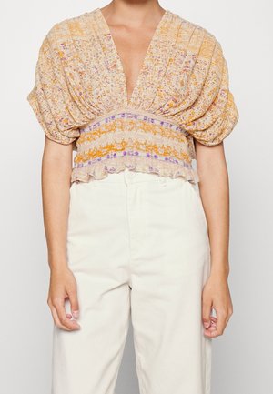 Vrouw draagt een beige, bloemenprint, geplooid cropped top met pofmouwen en witte, hooggehate broek tegen een effen achtergrond.