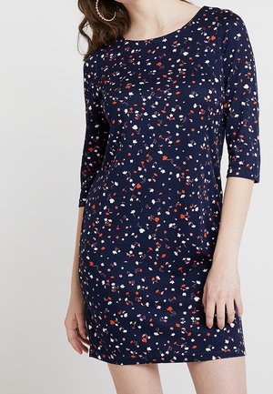 Marineblauwe jurk met rood, wit en roze bloemenpatroon, driekwart mouwen en een ronde halslijn gedragen door een staand persoon.