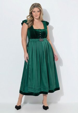 Mujer con vestido de terciopelo verde oscuro con mangas con volantes y una falda amplia plisada, zapatos negros puntiagudos, de pie sobre un fondo blanco.