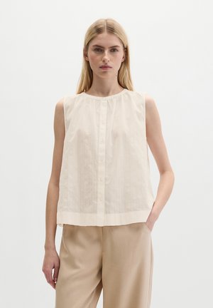 Femme blonde portant un chemisier blanc sans manches boutonné et un pantalon beige, debout avec une main dans la poche devant un fond uni.