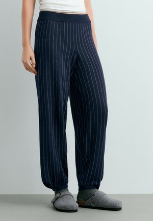 Personne portant un pantalon bleu marine à fines rayures, des chaussettes grises et des chaussons gris sans lacets, debout devant un fond clair uni.