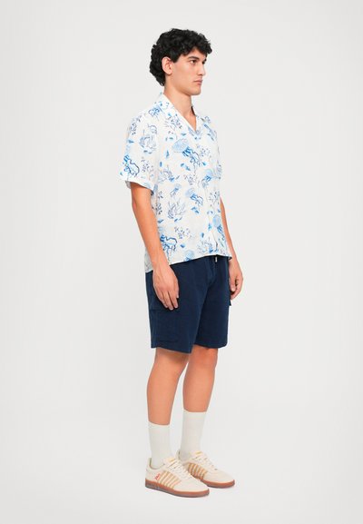 Camicetta bianca a maniche corte con stampa floreale blu, abbinata a pantaloni corti blu scuro. Sneakers con accenti beige e arancioni e calze chiare.
