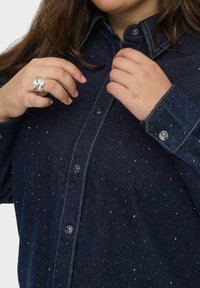 Person fastening dark blue denim shirt with small silver studs, wearing a large silver ring on left hand.