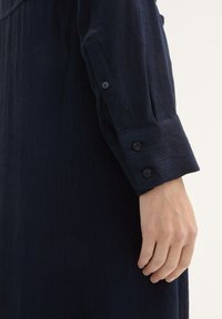 Maglietta a maniche lunghe a costine color blu navy con polsini con bottoni. Tessuto testurizzato, pieghe visibili e finitura liscia nella zona del colletto.