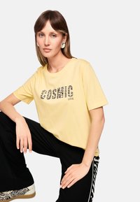 Yellow t-shirt features the text "COSMIC LOVE" in a zebra pattern. Model wears black pants with a zebra stripe design and white shoes.
