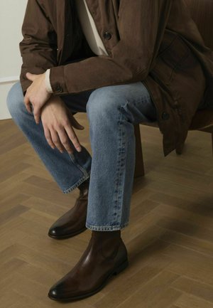 Stivaletti Chelsea in pelle marrone con punta a punta, indossati con jeans di denim blu chiaro e una giacca marrone, seduti su una sedia di legno.