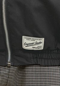 Veste noire avec fermeture éclair argentée et étiquette blanche rectangulaire portant la mention « Jack&Jones Creative Dept. Originals Studio Copenhagen 1990 » sur tissu pied-de-poule.