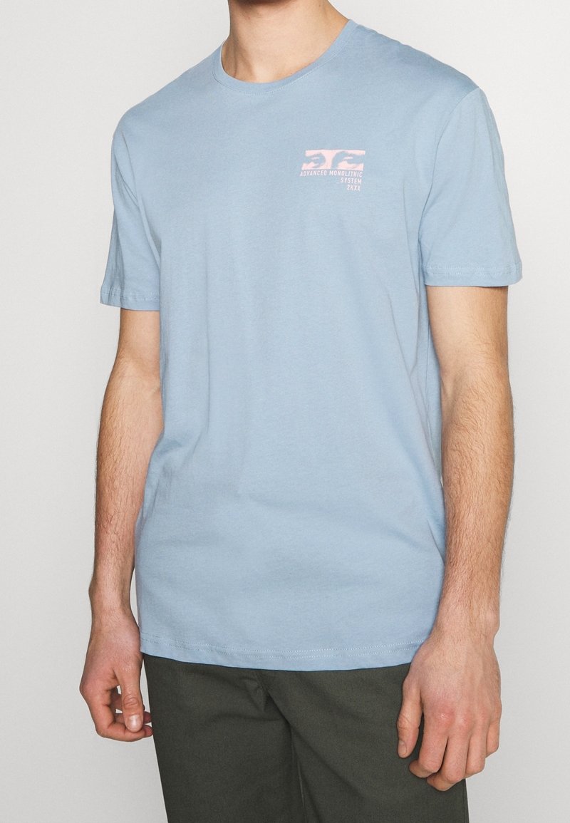 Homme portant un t-shirt bleu clair à manches courtes avec un petit graphisme rose et un texte sur la partie supérieure gauche de la poitrine, associé à un pantalon foncé.