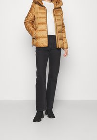 Brun oppustet jakke med hætte og lynlås, båret over en hvid strikket sweater, parret med højhalsede sorte jeans og sorte ankelstøvler.