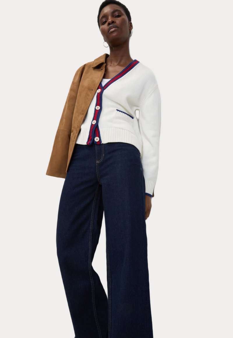 Cardigan bianco lavorato a maglia con accenti rossi e blu, e bottoni, indossato su jeans a gamba larga blu scuro. Il modello indossa anche una giacca color tan.
