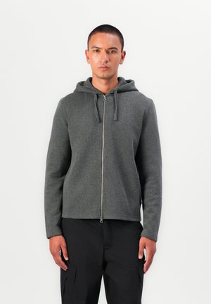 Gray zip-up hoodie with a hood, made of textured fabric. Features a front zipper and is worn with black cargo pants.