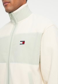 Fleecejakke i cremefarvet og lys grøn, med høj krave og logo-patch. Tekstureret stof med kontrasterende paneler og lynlås-lukning.