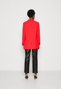 Personne debout de dos, portant un blazer rouge vif, un pantalon en simili cuir noir et des chaussures à talons noires, le tout sur un fond uni.