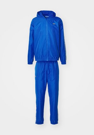 Tuta blu con giacca a zip con cappuccio e pantaloni con vita elastica. Realizzato in tessuto liscio con strisce laterali e un piccolo logo sul petto.