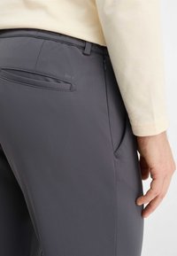 Grijze broek van glad, rekbaar materiaal met een achterzak, subtiele branding en een minimalistisch ontwerp. Zijzak zichtbaar.