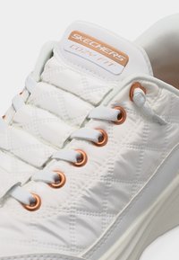 Λευκό αθλητικό παπούτσι Skechers με υφασμάτινη επένδυση καπιτονέ, ροζ-χρυσά μεταλλικά ζεμπόνια, λευκά κορδόνια και ετικέτα "Cozy Fit" στη μαξιλαροειδή γλώσσα.