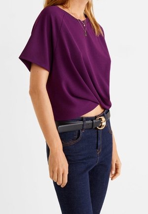 Woman wearing a short-sleeve purple top with a twisted front detail, paired with dark blue jeans and a black belt with a gold buckle.