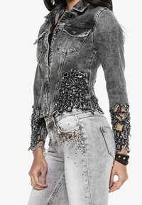 Harmaa farkkutakki, jossa on yksityiskohtaiset mustat pitsikoristeet ja helmikoristelut, klassisella kauluksella ja napillaisella etuosalla.