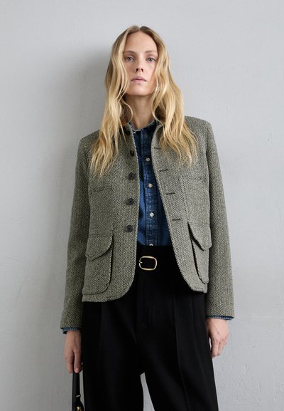 Sakó s vzorom houndstooth v šedohnedej farbe s golierom, štyrmi prednými vreckami a čiernymi gombíkmi, nosené cez džínsovú košeľu a čierne nohavice.