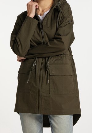 Personne portant une veste à capuche vert olive avec fermeture éclair à l'avant, cordons à la taille, grandes poches, et un jean bleu clair.