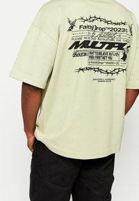 Camiseta verde clara de manga corta con estampados gráficos en negro, que incluyen texto y diseños de alambre de púa en la parte posterior. Ajuste casual.