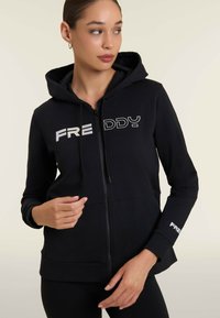 Sweat à zip noir en tissu doux avec poches avant et capuche. Présente le logo "FREDDY" en blanc sur la poitrine et la manche.