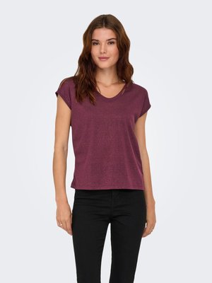 Femme aux cheveux bruns ondulés portant un haut violet à col en V à manches courtes et un pantalon noir, debout devant un fond clair uni.