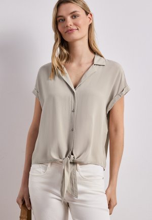 Junge Frau trägt ein beigefarbenes Kurzarmhemd, das an der Taille gebunden ist, und weiße Hosen und hält eine braune Tasche in der linken Hand.