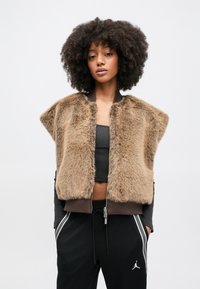Bruine faux bontvest met een cropped ontwerp, ritssluiting en geribbelde bruine zoom. Gedragen over een zwart cropped topje en gecombineerd met zwarte joggers.