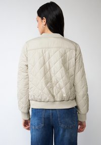 Blouson aviateur beige matelassé avec poignets et ourlet côtelés, présentant une texture lisse et un motif en losanges, associé à un jean en denim bleu.