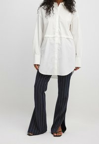 Chemise blanche à boutons avec un design court ; associée à un pantalon rayé bleu marine, présentant une jambe large et une fente en bas. Sandales noires.