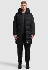 Black puffer coat with a hood, featuring a long design, rectangular quilted pattern, and a logo tag at the lower hem. Worn over dark trousers.