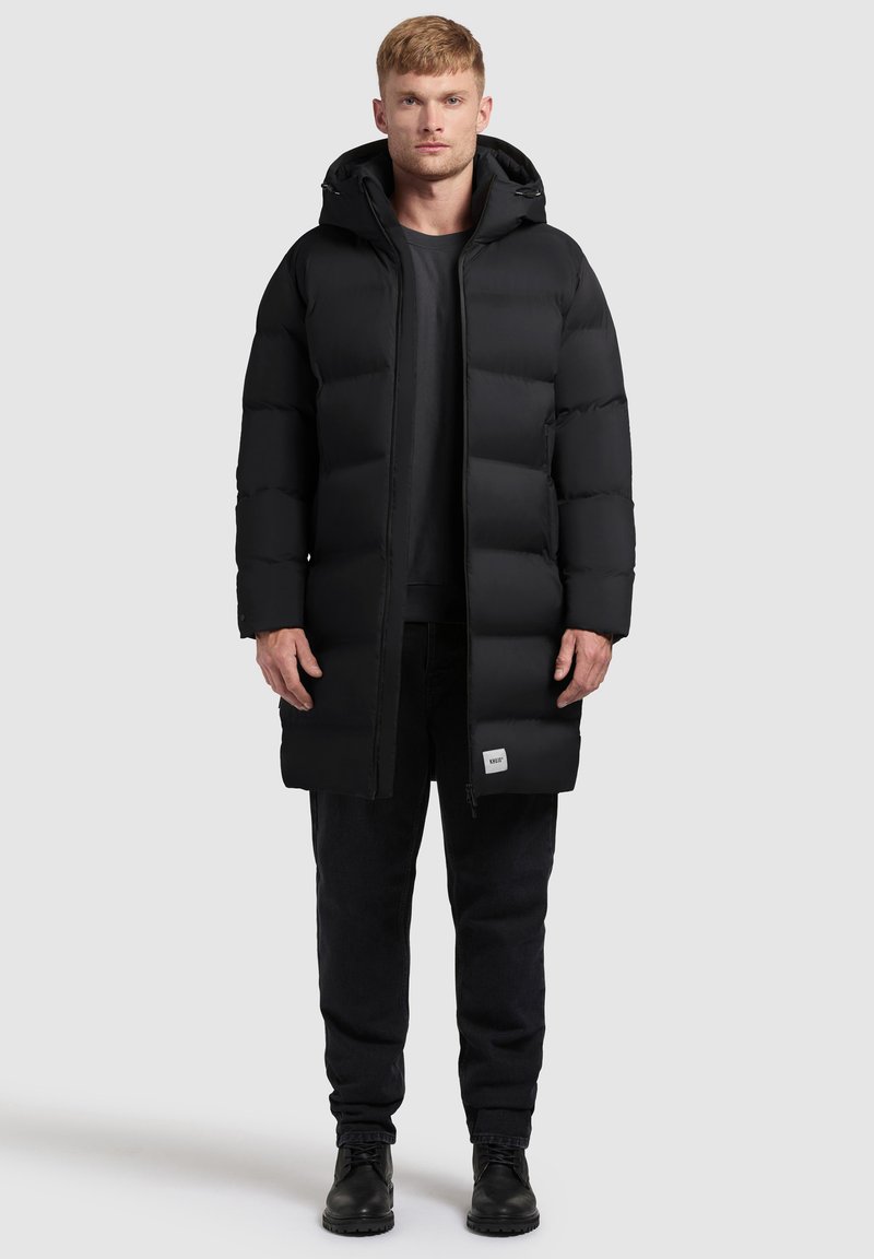 Black puffer coat with a hood, featuring a long design, rectangular quilted pattern, and a logo tag at the lower hem. Worn over dark trousers.
