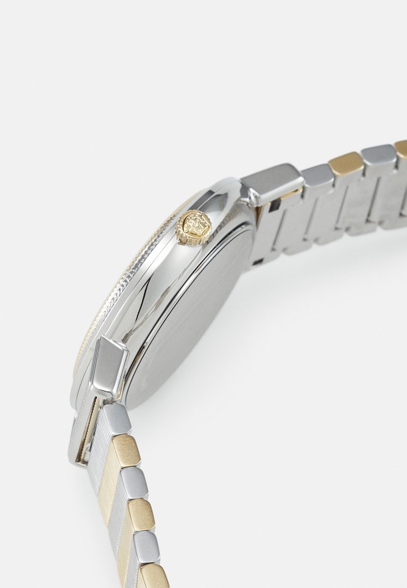 Silberne und goldfarbene Armbanduhr mit einem metallischen Armband, einem geriffelten silbernen Lünette und einer goldfarbenen Krone. Das Ziffernblatt der Uhr ist nicht sichtbar.