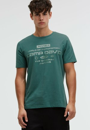 Young man with curly hair wearing a teal green Camp David motorsport graphic T-shirt and black pants, standing against a plain background.