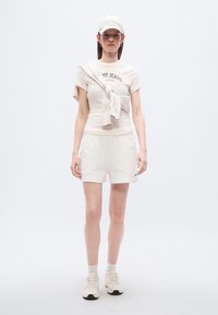 Κρεμ κοντομάνικη μπλούζα με το γραφικό «MY JEANS», συνδυασμένη με κρεμ σορτς. Ένα ελαφρύ κασκόλ είναι δεμένο γύρω από τη μπλούζα. Το σύνολο ολοκληρώνεται με ελαφριά αθλητικά παπούτσια και καπέλο.