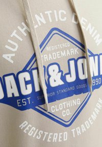 Beige hoodie med et blåt og hvidt logo-design og teksten "Jack & Jones", fremhævet af en snor og en glat stofstruktur.